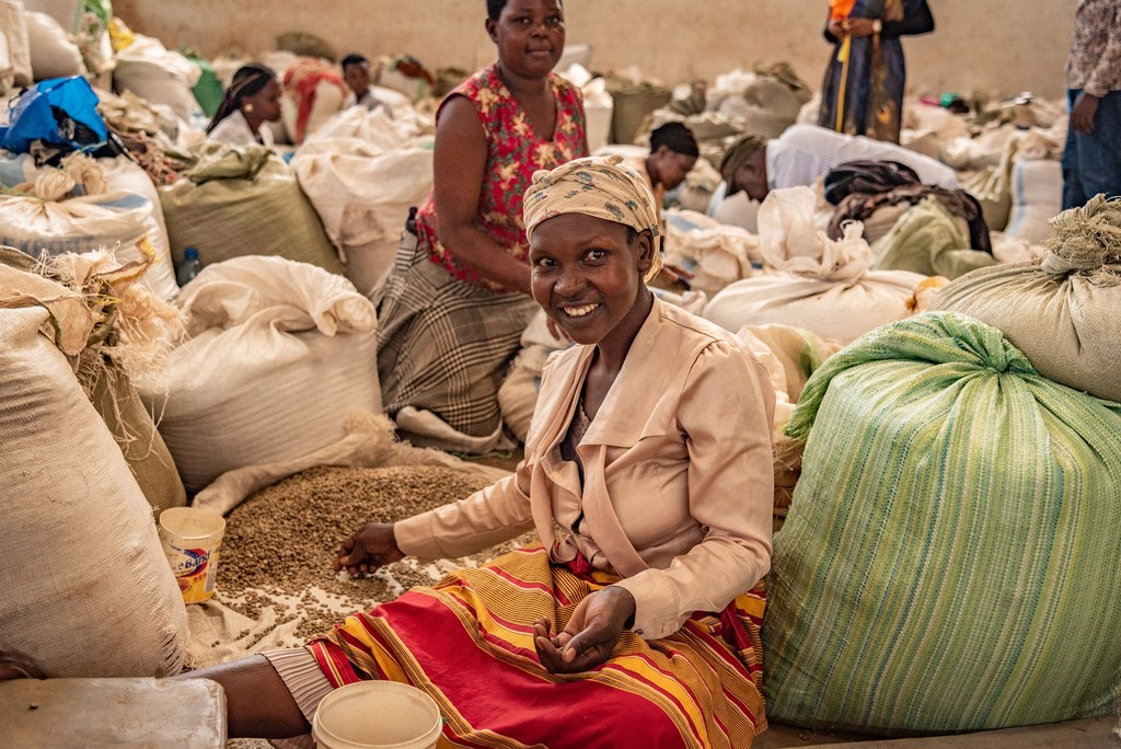 Sie sehen eine Arbeiterin der Fairtrade Bio Kaffee Kooperative aus Uganda. Die Frau sitz am Boden und sortiert die Bohnen mit der Hand aus. Um sie herum sind weiße Säcke voll Kaffee. Das Foto ist bei der Kaffee Reise 2019 nach Uganda, beim Besuch der Bio Fairtrade Kaffee Bauern und der Betriebsbesichtigung der Bio Fairtrade Kaffee Kooperative entstanden. Wir führen durch alle Schritte der Kaffeeernte und –Produktion.