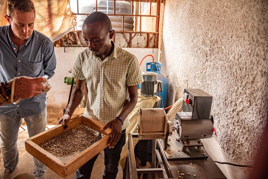 Im Bild prüft ein Mitarbeiter der Kooperative die Qualität des Kaffees mit einem großen Sieb, welches die Größe der Bohnen anzeigt. Daneben steht Thomas Angerer, der Reisebegleiter von life Earth Reisen. Das Foto ist bei der Kaffee Reise 2019 nach Uganda, beim Besuch der Bio Fairtrade Kaffee Bauern und der Betriebsbesichtigung der Bio Fairtrade Kaffee Kooperative entstanden. Wir führen durch alle Schritte der Kaffeeernte und –Produktion. Sie sehen die Qualitätskontrolle der Kaffeebohnen.